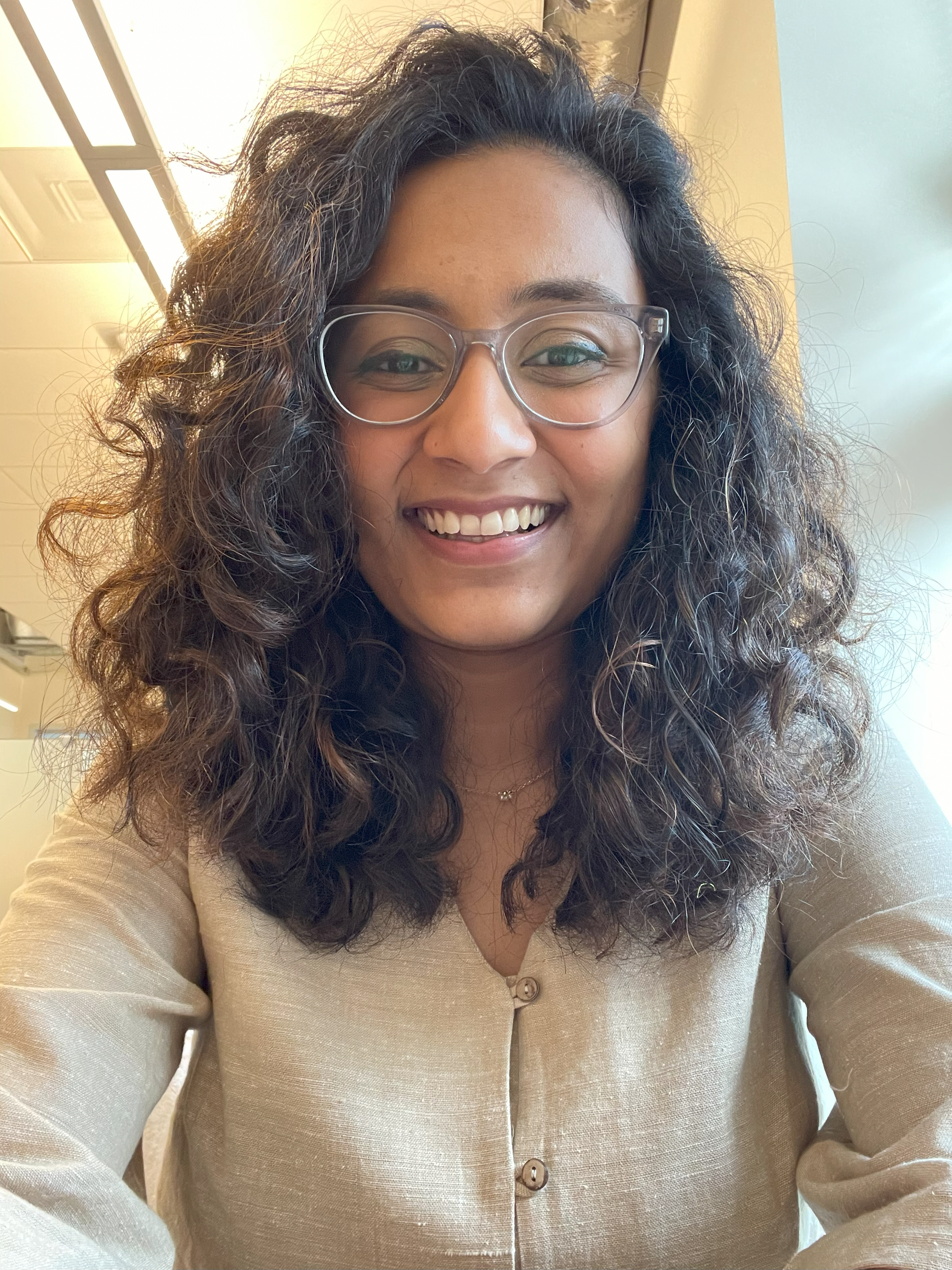 Photo of Rithi with beige button top, with shoulder-length curly brown hair and glasses