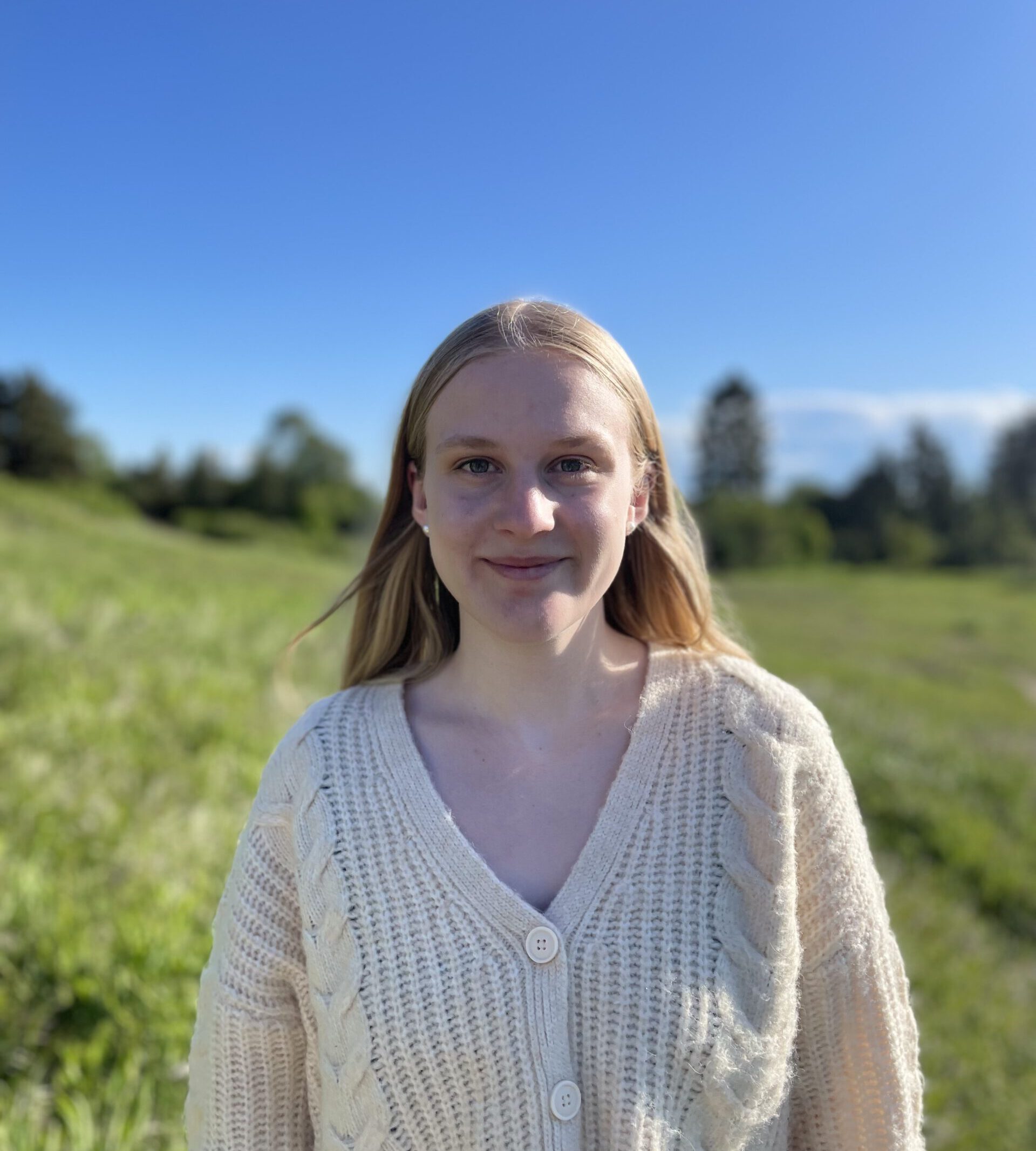 Photo of Sarah with white cardigan, blonde hair, with outdoors background of blue sky and green hill